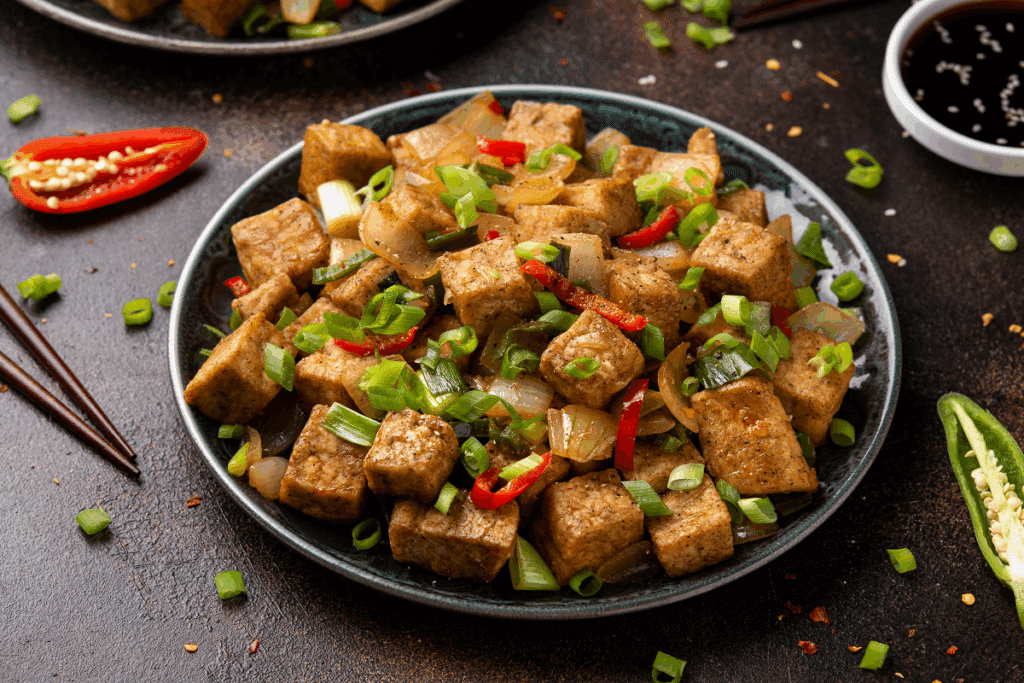 Prato de cerâmica escura com cubos dourados de tofu salteados com cebola e cebolinha, decorado com fatias de pimenta vermelha, pimentas ao redor e um recipiente com molho.
