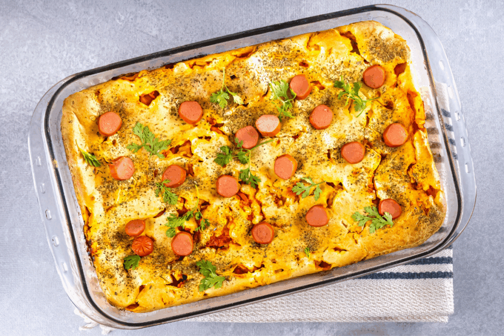 Vista superior de uma torta salgada assada em uma travessa retangular de vidro. A superfície dourada é decorada com rodelas de salsicha distribuídas por cima, polvilhada com ervas finas e finalizada com folhas frescas de coentro. A travessa está apoiada sobre um pano de prato claro com listras azuis.