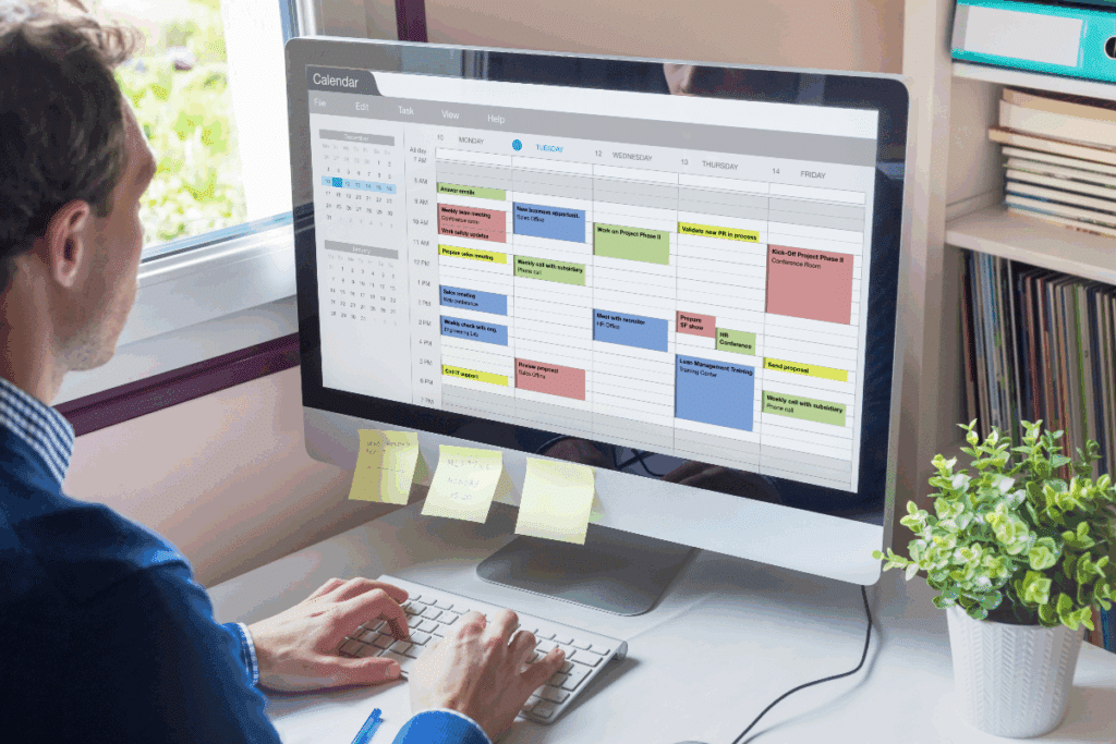 Imagem de um homem sentado à mesa de trabalho, utilizando um computador com uma agenda semanal digital aberta na tela. O calendário apresenta compromissos e tarefas organizados por dias e horários, destacados em diferentes cores. Abaixo do monitor, há pequenos lembretes adesivos. Sobre a mesa, aparecem um teclado, uma caneta azul e uma planta decorativa. Ao fundo, há uma janela e uma estante com livros e pastas.
