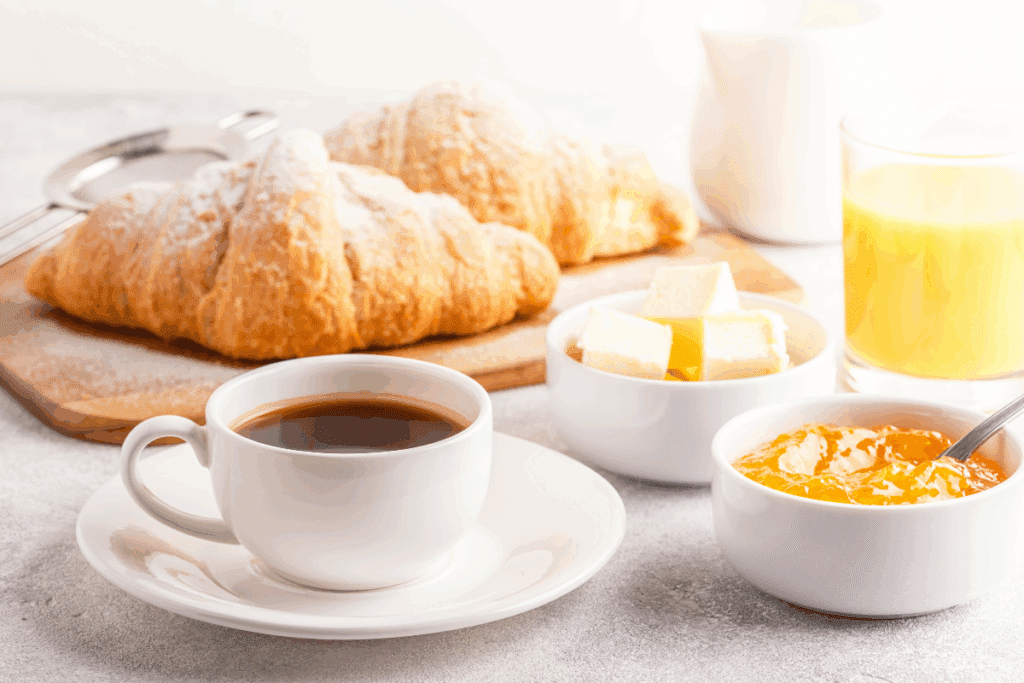 A imagem mostra uma mesa de café da manhã com dois croissants dourados sobre uma tábua de madeira, uma xícara branca com café preto, um copo de suco de laranja e uma jarra branca. Também há duas tigelas, uma com pedaços de manteiga e outra com geleia alaranjada e uma colher.