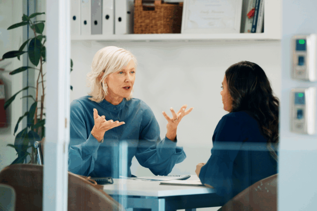 Duas mulheres sentadas próximas a uma mesa num escritório, onde uma mulher loira de blusa azul gesticula enquanto fala diretamente com a colega