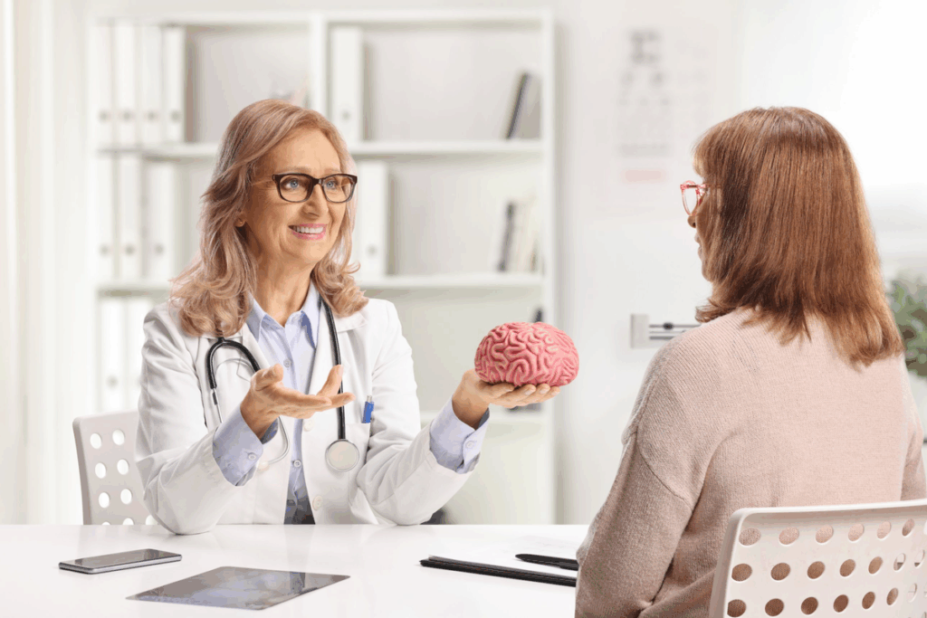 Psiquiatra sorridente de cabelos claros e óculos, com jaleco branco e estetoscópio, segurando um modelo de cérebro rosa enquanto conversa com uma paciente de cabelos castanhos vista de costas. As duas estão sentadas a uma mesa branca em um consultório claro, com estantes ao fundo.