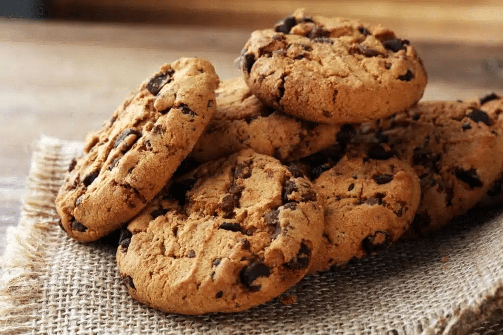 A imagem mostra vários cookies com pedaços de chocolate, empilhados sobre um tecido rústico. Os biscoitos apresentam aparência dourada e textura crocante, com gotas e pedaços de chocolate distribuídos pela massa