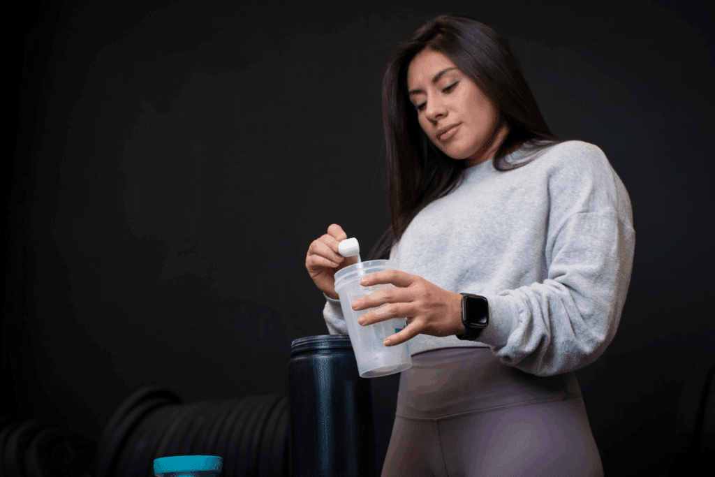 Mulher jovem de cabelos escuros, moletom cinza e legging, colocando uma dose de creatina em pó dentro de uma coqueteleira transparente, em um ambiente de academia com fundo escuro.