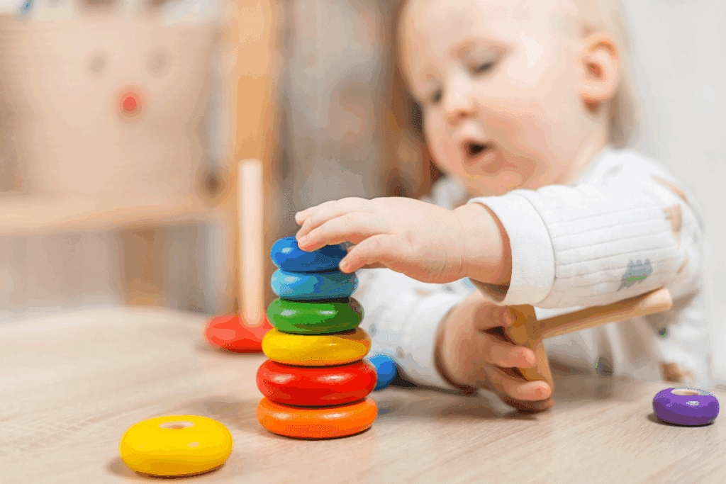 A imagem mostra uma criança pequena brincando com um brinquedo educativo de encaixe. Sobre uma mesa de madeira, ela empilha peças circulares coloridas em uma haste, formando uma sequência com tons de laranja, vermelho, amarelo, verde e azul. Outras peças aparecem espalhadas pela superfície.