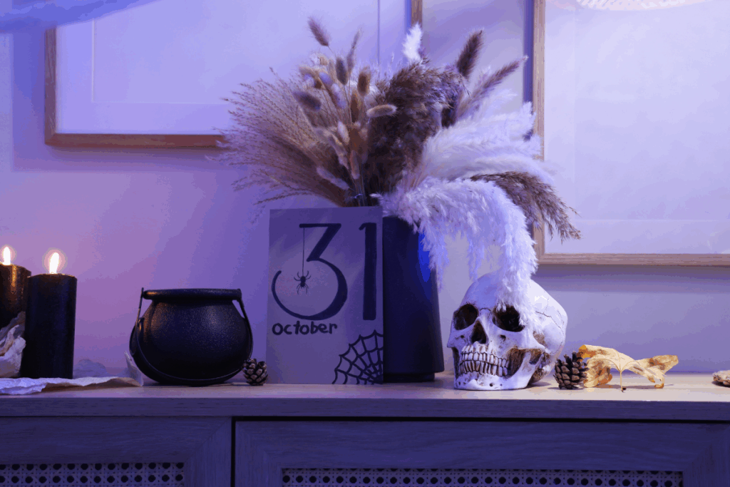 Decoração de Halloween sobre um aparador de madeira escura, iluminada por luzes em tons de roxo e azul. À esquerda, há duas velas pretas acesas e um pequeno caldeirão preto. No centro, um calendário decorativo exibe o número 31, a palavra “october”, uma aranha e uma teia desenhadas em preto. Ao lado, há um vaso alto e escuro com folhagens secas e plumas em tons de bege, marrom e branco. À direita, um crânio decorativo branco está acompanhado de pinhas e folhas secas amareladas. Ao fundo, há uma parede clara com quadros de moldura de madeira.