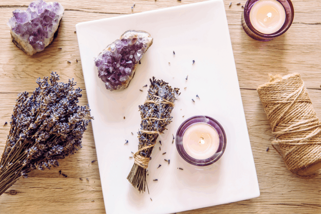 Mesa de madeira disposta com elementos rituais, incluindo um maço de lavanda seca atado com corda, drusas de ametista roxa, velas acesas e um rolo de fio de juta