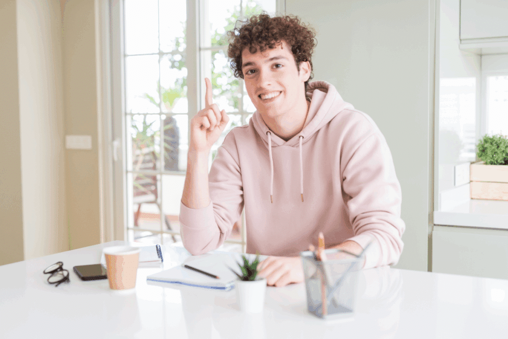 A imagem mostra um jovem estudante sentado à mesa em um ambiente claro e organizado. Ele sorri para a câmera enquanto mantém um dos dedos levantado, em um gesto que sugere uma ideia ou descoberta. Sobre a mesa, aparecem cadernos, lápis, óculos, celular, copo e um porta-lápis.
