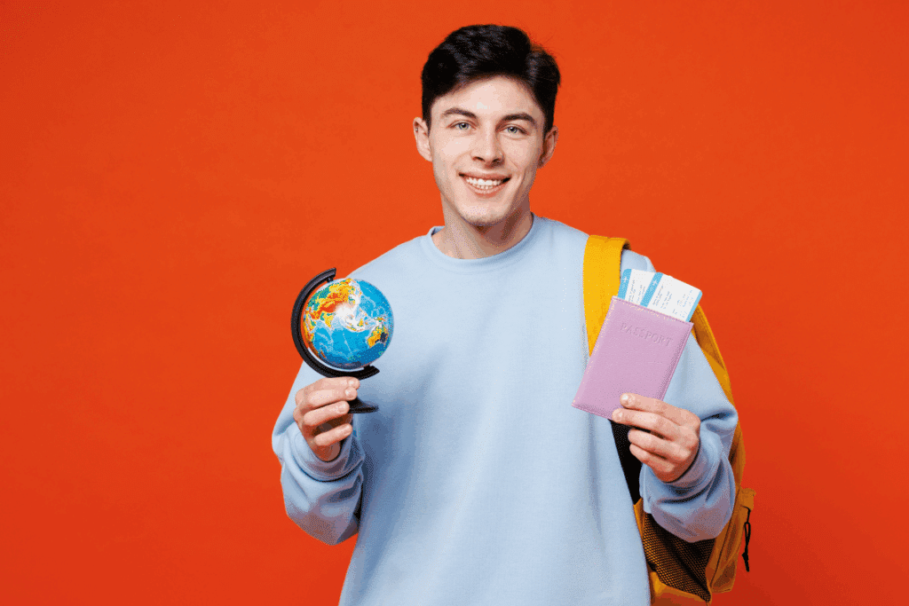 Fotografia de um jovem de cabelos curtos, lisos e pretos, sorrindo e olhando diretamente para a câmera. Ele veste um moletom azul-claro e carrega uma mochila amarela nos ombros. Na mão esquerda, segura um pequeno globo terrestre colorido, com suporte preto. Na mão direita, exibe um passaporte rosa com a palavra “PASSPORT” gravada na capa e cartões de embarque ou documentos de viagem parcialmente visíveis na parte superior em fundo vermelho.
