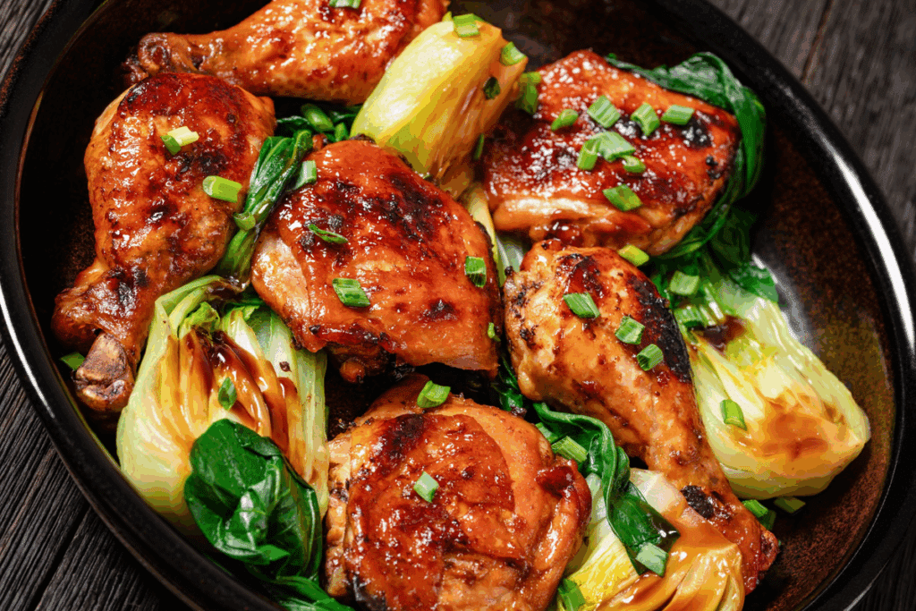 Travessa escura com coxas e sobrecoxas de frango glaceado e douradas, acompanhadas de acelga chinesa e cebolinha picada