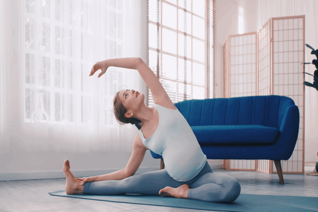 A imagem mostra uma mulher grávida praticando yoga em casa, sentada sobre um tapete azul. Ela realiza um exercício de alongamento lateral, mantendo uma perna estendida, a outra dobrada e um dos braços elevado sobre a cabeça, enquanto a mão oposta segura a perna estendida. A gestante veste uma regata azul-clara e uma legging azul-acinzentada.