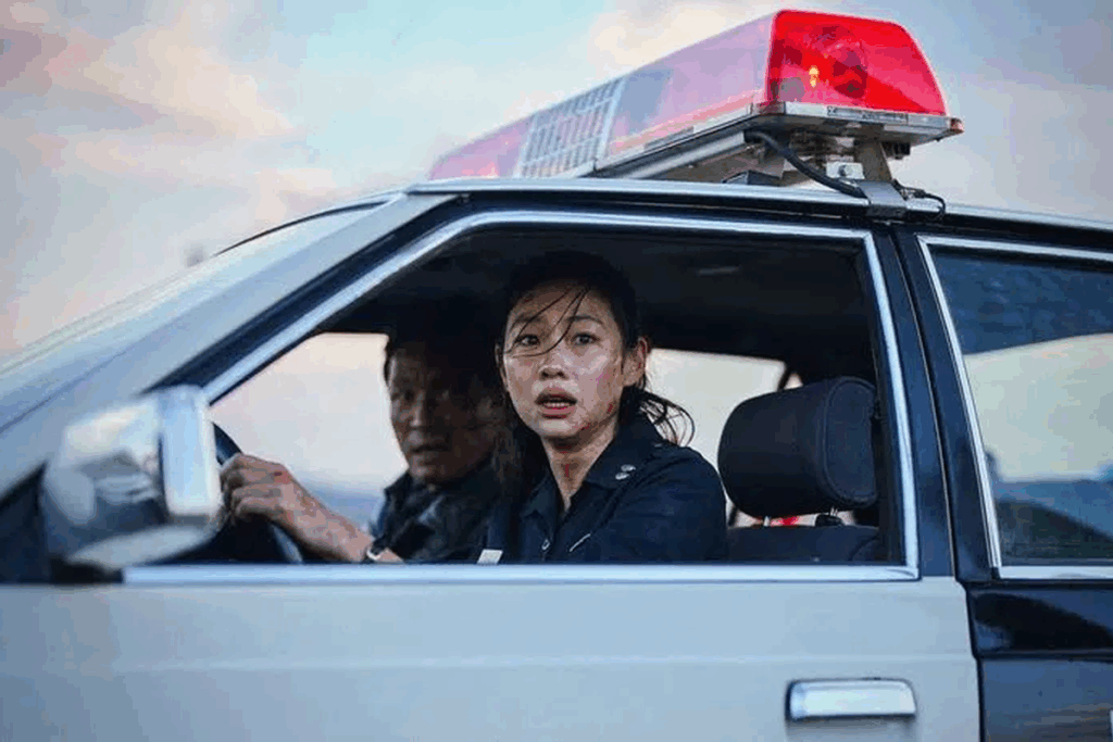 Cena do filme "Hope" em que uma mulher de cabelos escuros, com o rosto sujo e expressão assustada, olha pela janela de uma viatura policial. Ao lado dela, um homem dirige. No teto do carro, há uma sirene vermelha, sob um céu de fim de tarde.