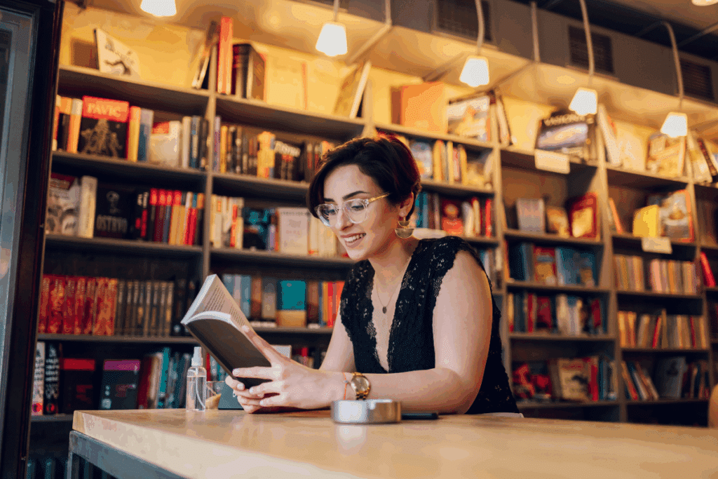 Uma mulher de cabelos curtos e escuros, usando óculos, está sentada à mesa e lê um livro aberto. Ela sorri enquanto acompanha a leitura. Ao fundo, há uma grande estante repleta de livros de diferentes tamanhos e cores, iluminada por luminárias pendentes de luz quente. A cena transmite um ambiente acolhedor de biblioteca ou livraria.