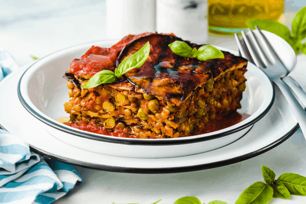 Fotografia de uma porção quadrada de lasanha vegetariana servida em um prato branco com bordas finas escuras, sobre uma superfície clara. O prato apresenta camadas de legumes e pequenos grãos, intercaladas com molho vermelho de tomate. A parte superior está dourada e coberta parcialmente pelo molho, com folhas verdes frescas de manjericão como decoração. À direita, há um garfo e uma faca de metal, enquanto um pano de cozinha azul-claro e branco aparece dobrado no lado esquerdo. Ao fundo, aparecem folhas de manjericão e um recipiente de vidro com azeite.