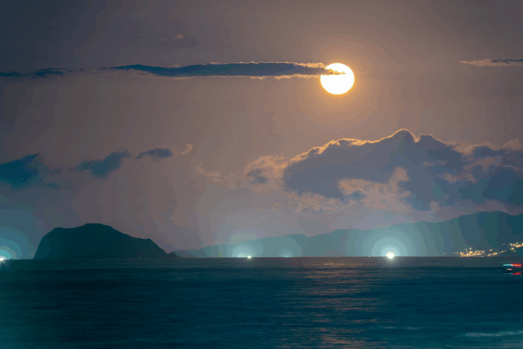 Lua cheia alaranjada sobre o mar, com montanhas ao fundo e luzes de barcos refletidas na água.