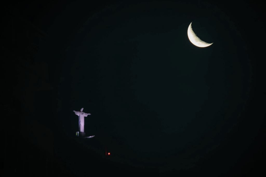 Céu noturno no Rio de Janeiro com a Lua em formato de foice no alto e, mais abaixo, a estátua do Cristo Redentor iluminada, de braços abertos.