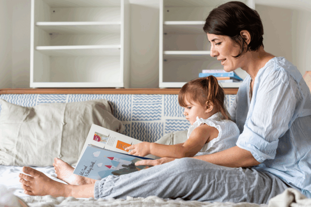 Fotografia de uma mulher de cabelos curtos e escuros sentada em um sofá com uma menina pequena no colo. As duas leem juntas um livro infantil ilustrado com um castelo, e a menina aponta para a página. Ao fundo há uma estante branca com livros coloridos.