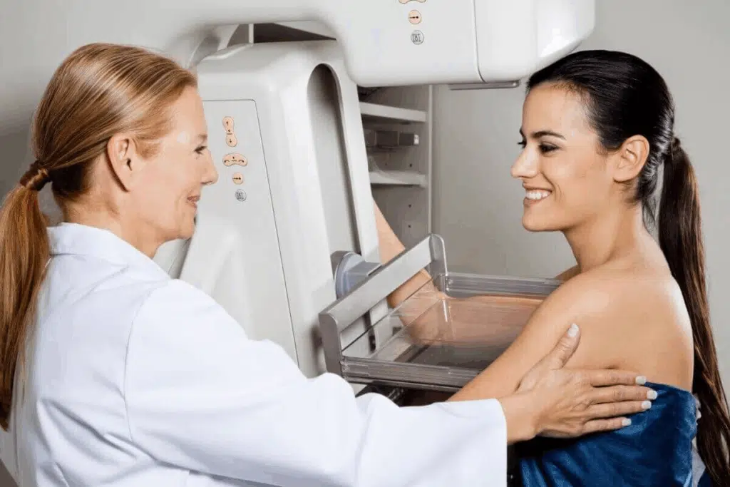 Uma médica loira, vestindo um jaleco branco, realiza um exame de mamografia em uma paciente de cabelos escuros presos em um rabo de cavalo. Ambas estão de perfil, sorrindo uma para a outra, transmitindo uma sensação de tranquilidade e cuidado. A paciente está posicionada em frente ao mamógrafo branco, com a mama apoiada na plataforma de compressão do aparelho. A médica segura delicadamente o braço da paciente para auxiliá-la no posicionamento correto.