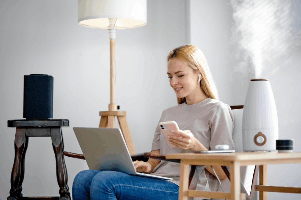 Mulher sentada em uma poltrona usa laptop e celular ao lado de um umidificador branco que solta vapor no ambiente.