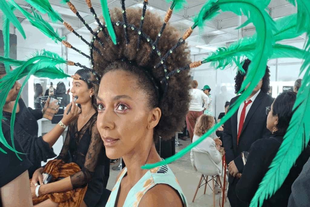 Imagem de uma modelo com cabelos crespos e volumosos, usando um penteado elaborado com tranças finas, aplicações brilhantes e longas penas verdes que se projetam em diferentes direções. Ela veste uma roupa em tons claros e apresenta maquiagem com destaque para os olhos em tons rosados.