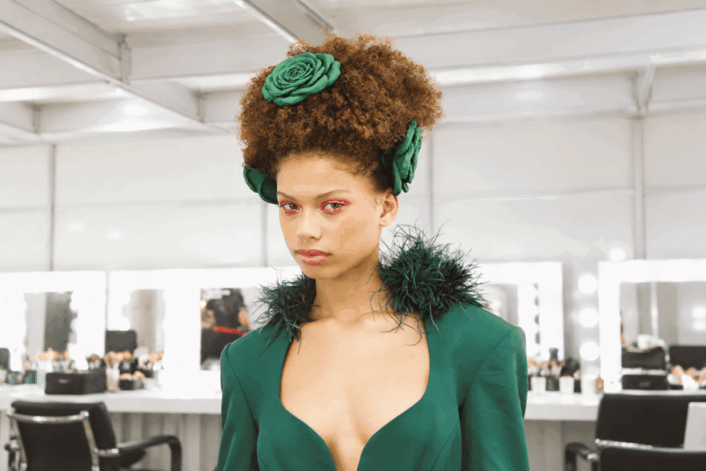 Imagem de uma modelo com cabelos castanhos, cacheados e volumosos, presos em um penteado alto e decorados com flores verdes. Ela veste uma roupa verde-escura com decote profundo e detalhes de plumas nos ombros. A maquiagem destaca os olhos com sombra rosa vibrante. Ao fundo, aparecem espelhos iluminados, cadeiras e produtos de maquiagem em um camarim.
