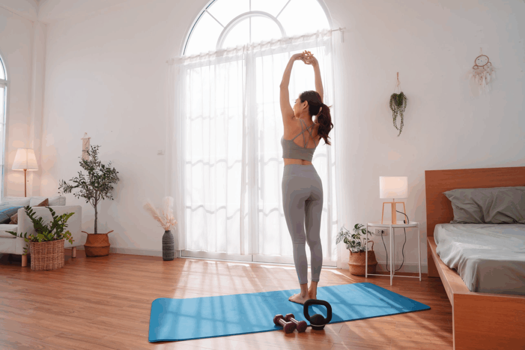 Fotografia de uma mulher de costas, com top e legging cinza, se alongando com os braços erguidos sobre um tapete de ioga azul, diante de uma grande janela em arco com cortinas brancas. No chão, há halteres e um kettlebell, e o quarto é claro, com cama, plantas e luminárias.