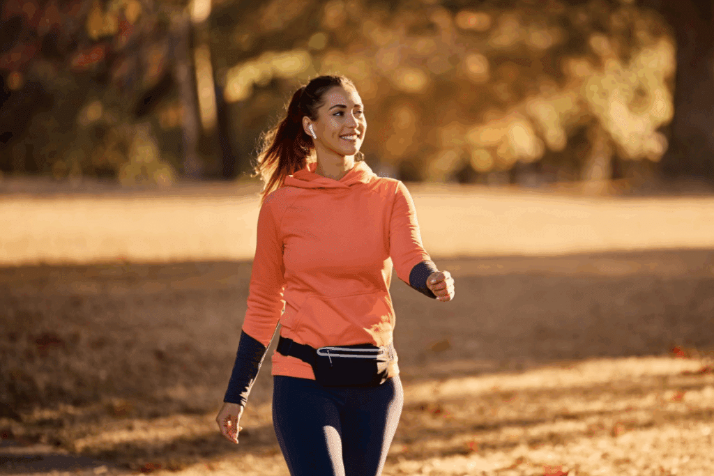 Mulher de cabelos castanhos presos em um rabo de cavalo caminha sorrindo em um parque arborizado, durante o fim da tarde. Ela usa fones de ouvido brancos, blusa com capuz na cor coral, com mangas azul-marinho, calça legging azul-escura e pochete preta na cintura. Ao fundo, há árvores com folhagem em tons de verde, amarelo e marrom, além de um caminho coberto por folhas secas. A luz dourada do sol ilumina a cena, criando uma atmosfera acolhedora.