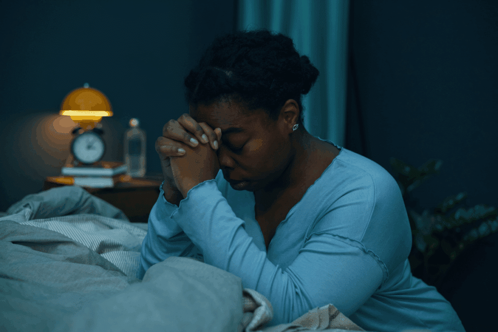 Fotografia de uma mulher negra, com cabelos pretos e presos em um coque, sentada na cama em um quarto com iluminação baixa e tons azulados. Ela veste uma blusa azul de mangas compridas, usa pequenos brincos prateados e mantém os olhos fechados, com as mãos entrelaçadas diante do rosto e a testa apoiada nos dedos, em uma postura de oração ou meditação. À esquerda, sobre um criado-mudo, há um relógio despertador com cúpula amarela iluminada, alguns livros e uma garrafa transparente. A cama está coberta por roupas de cama em tons de cinza, enquanto o fundo escuro e a luz suave criam uma atmosfera noturna e silenciosa.