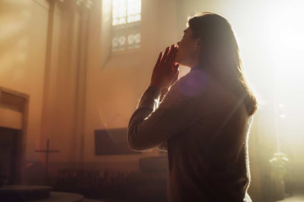 Mulher ajoelhada em uma igreja, rezando e sendo iluminada pela luz do sol