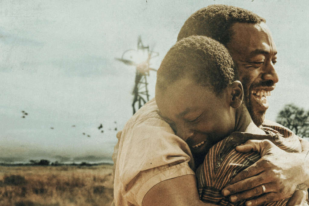 Cena do filme O Menino que Descobriu o Vento: um pai, sorrindo, abraça o filho em um campo seco. Ao fundo há um moinho de vento feito à mão e pássaros no céu.