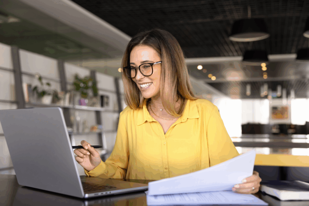Uma mulher sorridente usando óculos e blusa amarela trabalha em um notebook enquanto segura papéis em um escritório