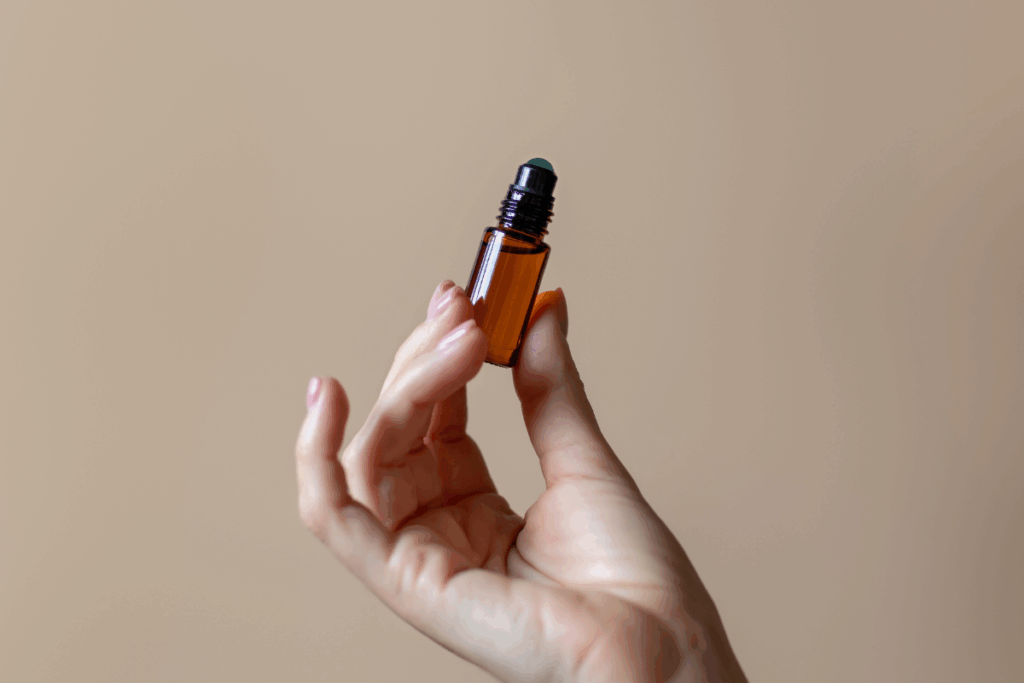 Um close das mãos de uma pessoa segurando um pequeno frasco de vidro âmbar com aplicador roll-on, contra um fundo bege-claro