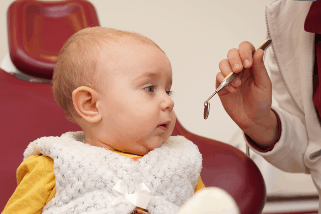 Um bebê loiro, vestindo um colete felpudo branco, está sentado em uma cadeira de dentista vermelha, olhando curioso para um espelho clínico que um profissional segura próximo ao seu rosto