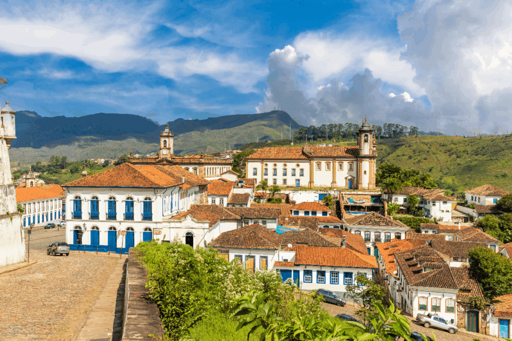 Vista panorâmica do centro histórico de Ouro Preto, em Minas Gerais, com casarões coloniais de fachadas brancas, portas e janelas coloridas e telhados de barro. Ao fundo, destacam-se igrejas históricas com torres imponentes, cercadas por montanhas verdes. O céu azul, com grandes nuvens brancas, completa a paisagem da cidade histórica.