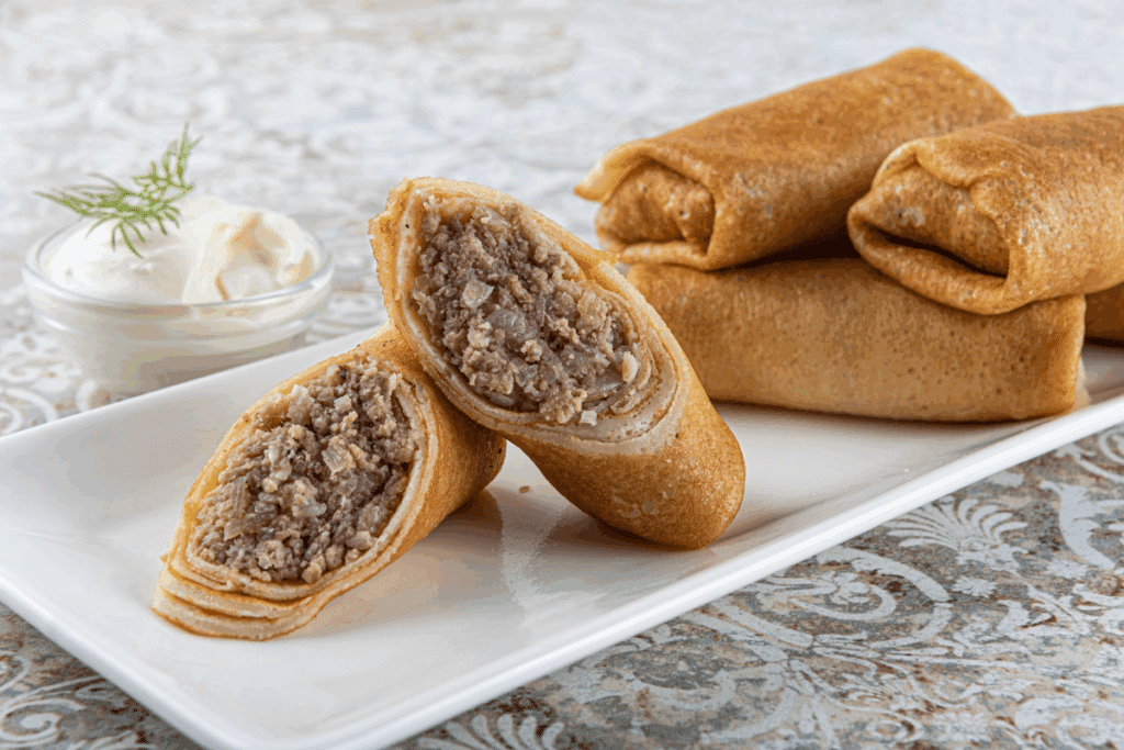 Panquecas enroladas recheadas com carne moída, uma delas cortada ao meio mostrando o recheio, servidas com um pote de creme e endro em um prato branco