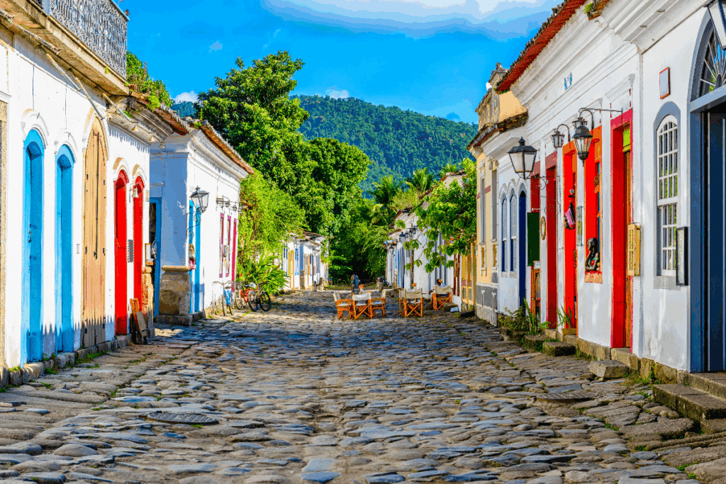 Rua de pedras do centro histórico de Paraty, no Rio de Janeiro, cercada por casarões coloniais de fachadas brancas, portas e janelas coloridas. Ao longo da rua, há árvores, bicicletas e mesas com cadeiras de madeira. Ao fundo, destacam-se montanhas cobertas por vegetação verde sob um céu azul.