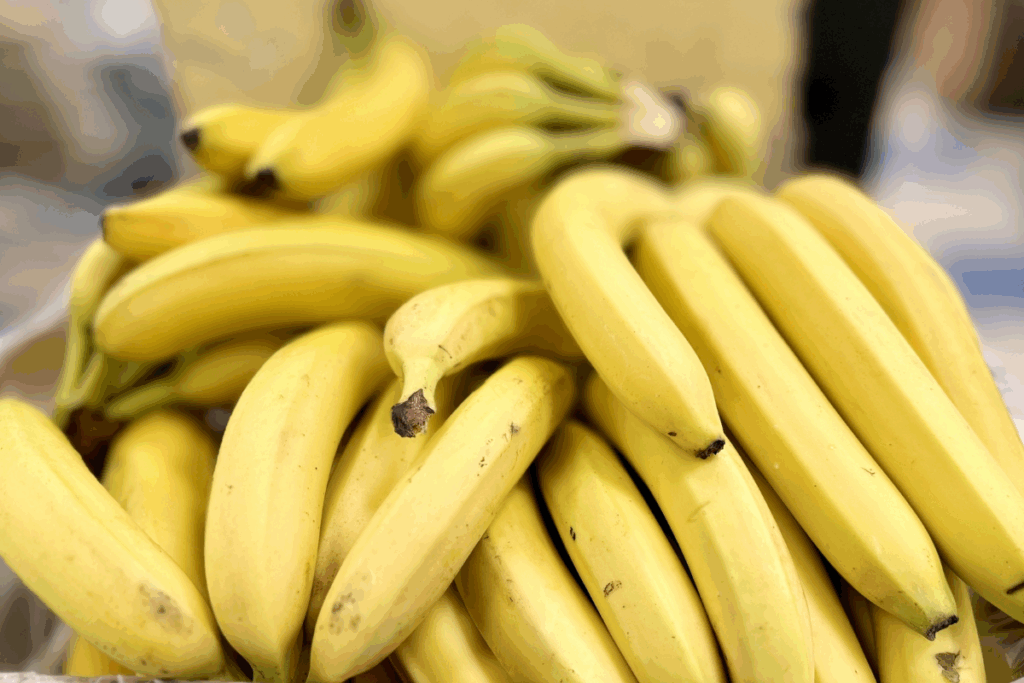 Fotografia em close-up de um grande agrupamento de bananas maduras, com cascas amarelas, algumas pequenas manchas escuras e pontas amarronzadas. As frutas estão sobrepostas e ocupam quase toda a imagem, formando uma composição volumosa. As bananas em primeiro plano aparecem mais nítidas, destacando a textura das cascas, enquanto as frutas ao fundo ficam desfocadas. A iluminação ressalta os tons amarelos e cria uma aparência natural, com o foco concentrado nas bananas.