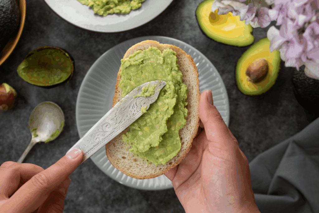 Foto do preparo de uma torrada com abacate. Em destaque no centro, mãos seguram uma fatia de pão de forma rústico enquanto espalham uma pasta de abacate verde-clara sobre ela com uma faca de metal trabalhada. O pão está posicionado sobre um prato raso cinza claro. No fundo, sobre uma superfície escura de textura mista, veem-se metades de abacate, uma colher, a casca vazia da fruta e flores em tons suaves de rosa e lilás ao canto superior direito