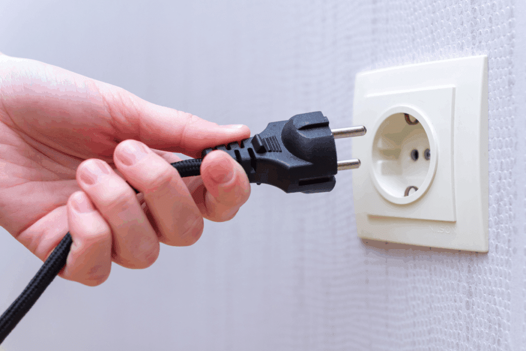 Mão segurando um plugue elétrico preto de dois pinos, prestes a ser inserido ou removido de uma tomada branca instalada em uma parede clara e levemente texturizada