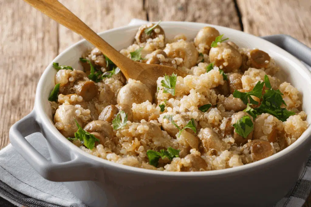 Prato de quinoa com cogumelos salteados, servido em uma travessa branca e decorado com folhas de salsa. Uma colher de madeira repousa sobre a preparação.