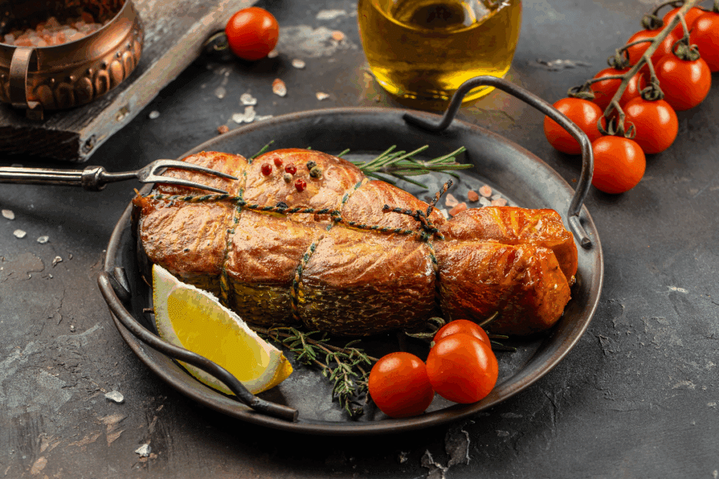 A imagem mostra um peixe assado ou grelhado, preparado inteiro em uma peça grossa e amarrado com barbante culinário. A superfície está bem dourada, com marcas de cocção e temperos aparentes, incluindo pimentas e ervas aromáticas, como alecrim e tomilho. O peixe é servido em uma frigideira rústica, acompanhado de tomates-cereja e uma fatia de limão.