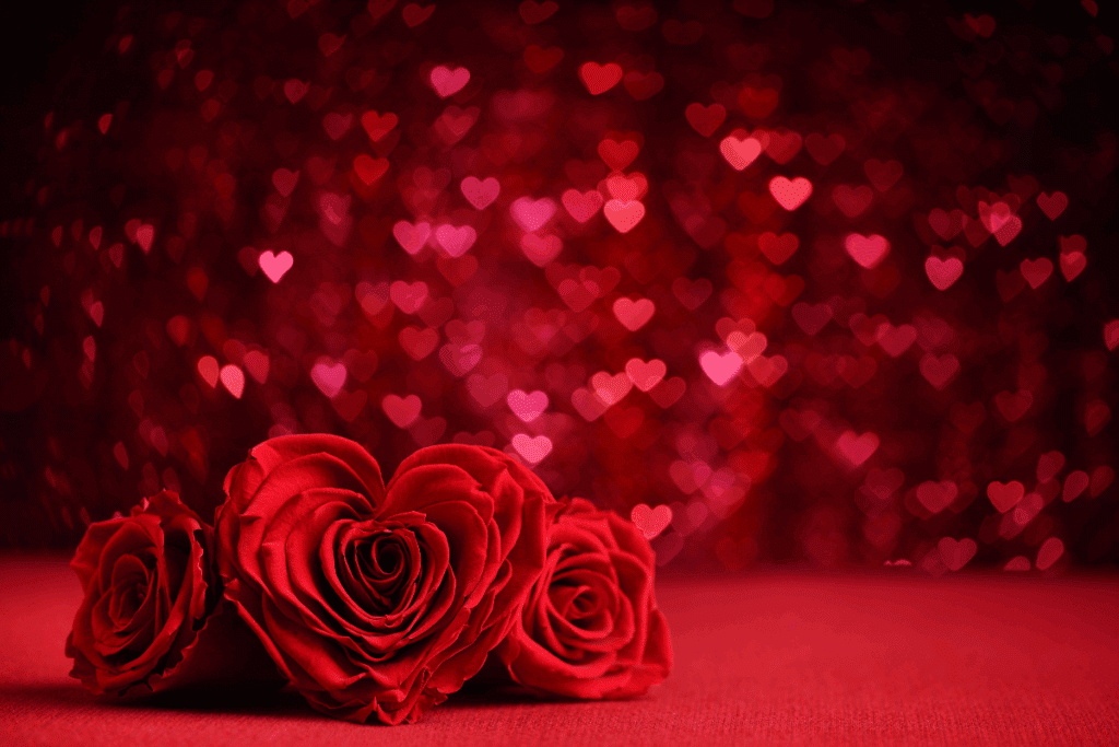 A imagem mostra rosas vermelhas sobre uma superfície também vermelha, em uma composição romântica. Ao centro, uma das rosas tem as pétalas organizadas de maneira que lembram o formato de um coração. Ao fundo, diversos pontos luminosos desfocados em forma de coração, em tons de vermelho e rosa