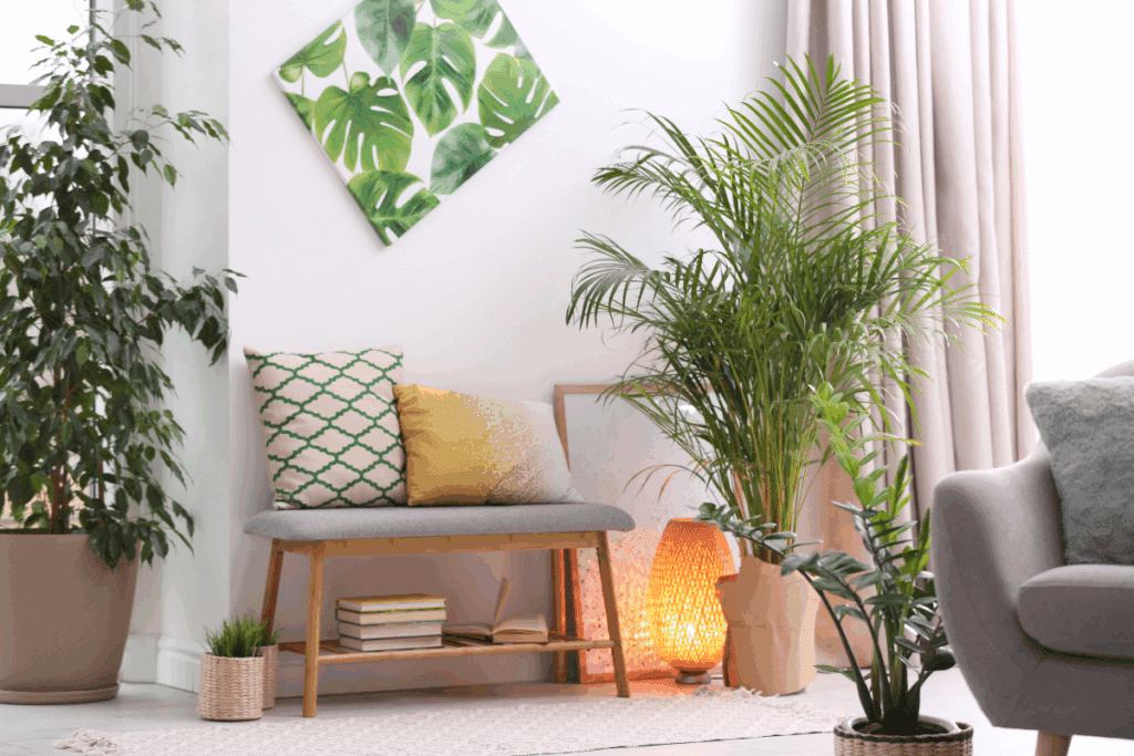 Canto de sala decorado com um banco de madeira clara e assento cinza, acompanhado de almofadas brancas com estampa verde e amarelo-mostarda. Acima, há um quadro inclinado com folhas verdes grandes sobre fundo branco. O ambiente possui várias plantas ornamentais em vasos de diferentes tamanhos, incluindo uma palmeira alta à direita e uma planta de folhas largas à esquerda. Sob o banco, há livros empilhados e um livro aberto. Ao lado, uma luminária de chão com cúpula rendada em tom alaranjado emite luz quente. O espaço tem paredes brancas, cortinas bege-rosadas e tapete claro.