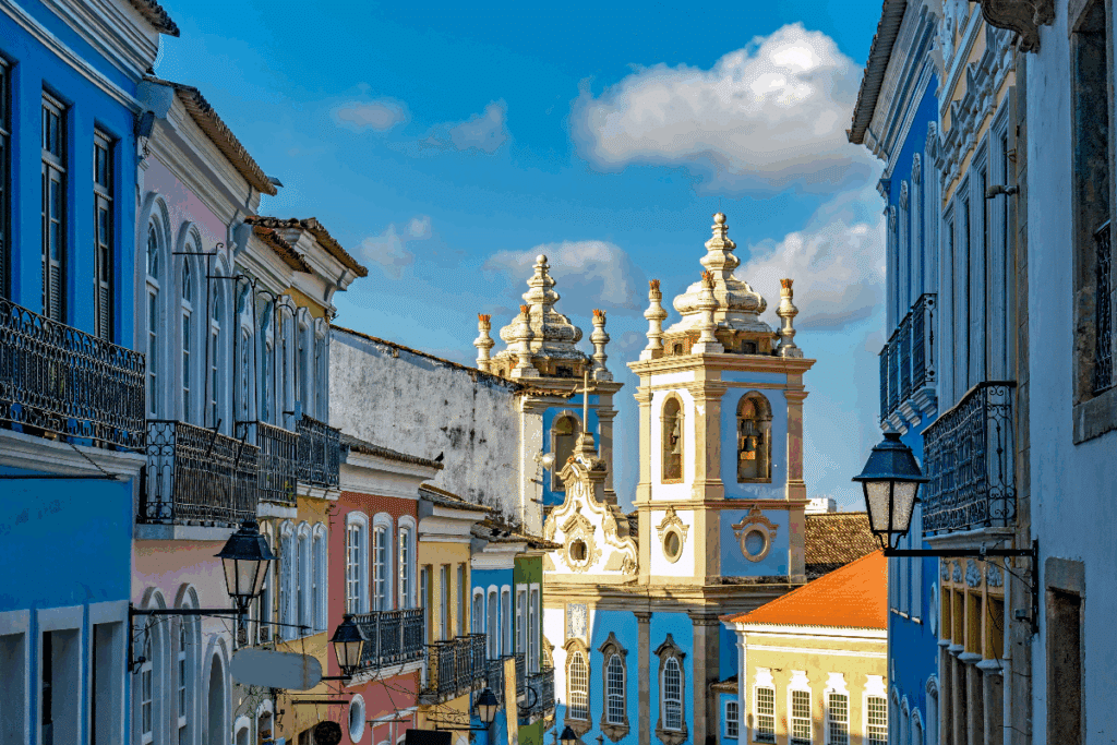 Vista de uma rua do centro histórico de Salvador, na Bahia, com casarões coloniais de fachadas azuis, rosas e amarelas, janelas altas e sacadas de ferro. Ao fundo, destacam-se as torres ornamentadas de uma igreja histórica, com detalhes arquitetônicos em tons claros. O céu azul, com nuvens brancas, completa a paisagem.