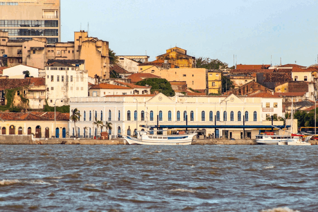 Vista panorâmica do centro histórico de São Luís, no Maranhão, com casarões coloniais, construções antigas e edifícios de diferentes alturas. Em primeiro plano, destaca-se um grande prédio histórico de fachada clara, com portas e janelas azuis, localizado à beira-mar. Algumas embarcações estão atracadas próximo à orla, enquanto as águas ocupam a parte inferior da imagem.