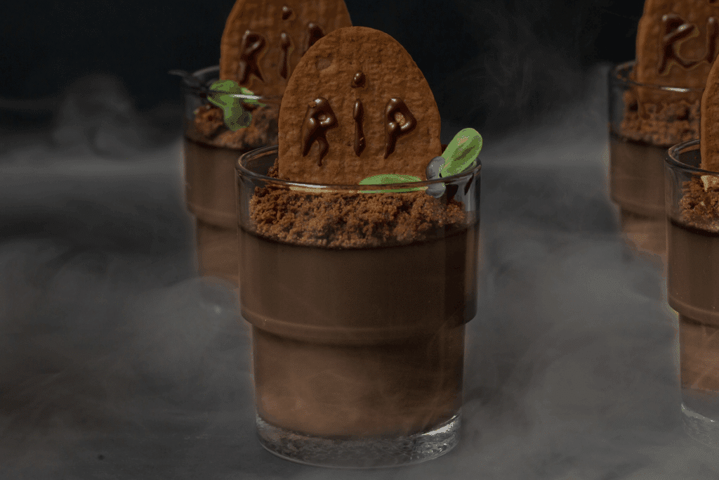 Sobremesa de Halloween em um copo de vidro, decorada como um pequeno túmulo, em destaque no centro, com outros copos iguais desfocados ao fundo. O copo tem uma camada de creme de chocolate claro, coberta por cacau em pó imitando terra, com um biscoito de chocolate em formato de lápide escrito "RIP" com calda de chocolate. Folhinhas verdes enfeitam a terra, e uma névoa branca se espalha ao redor, sobre um fundo preto.