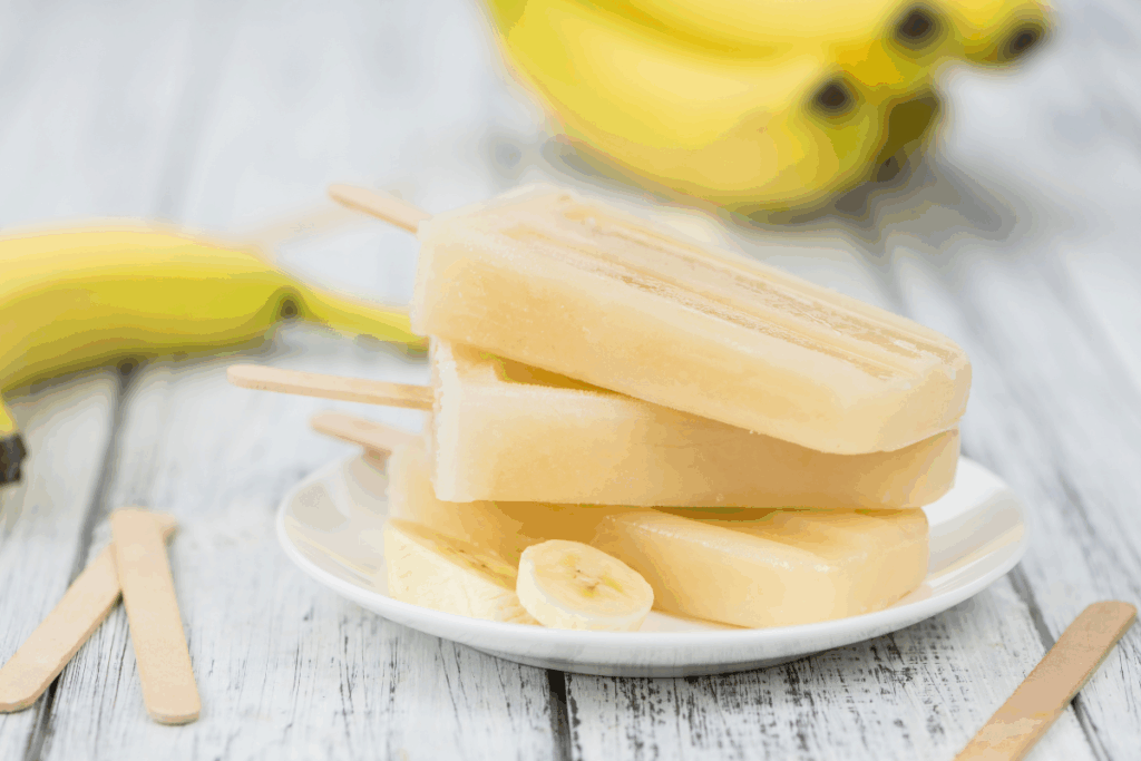 Picolés caseiros de banana empilhados em um pequeno prato branco sobre uma mesa de madeira clara, acompanhados por rodelas e cachos da fruta ao fundo