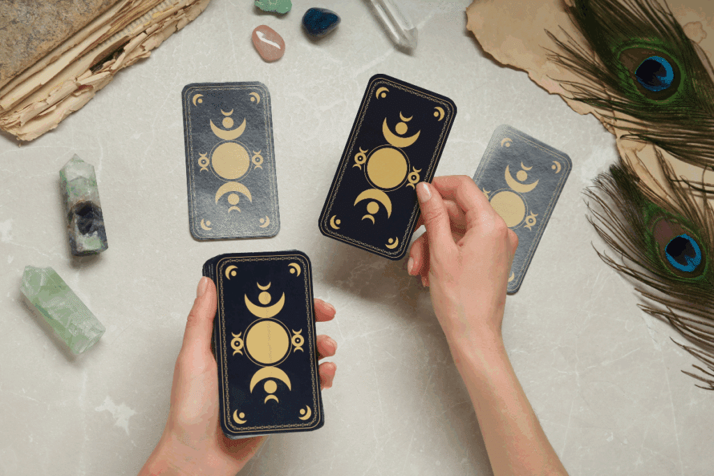 A imagem mostra uma pessoa manuseando cartas de tarô sobre uma mesa clara. As cartas têm fundo escuro e detalhes dourados com símbolos de luas e círculos. Ao redor, aparecem cristais de diferentes cores, penas de pavão, papéis envelhecidos e outros elementos decorativos