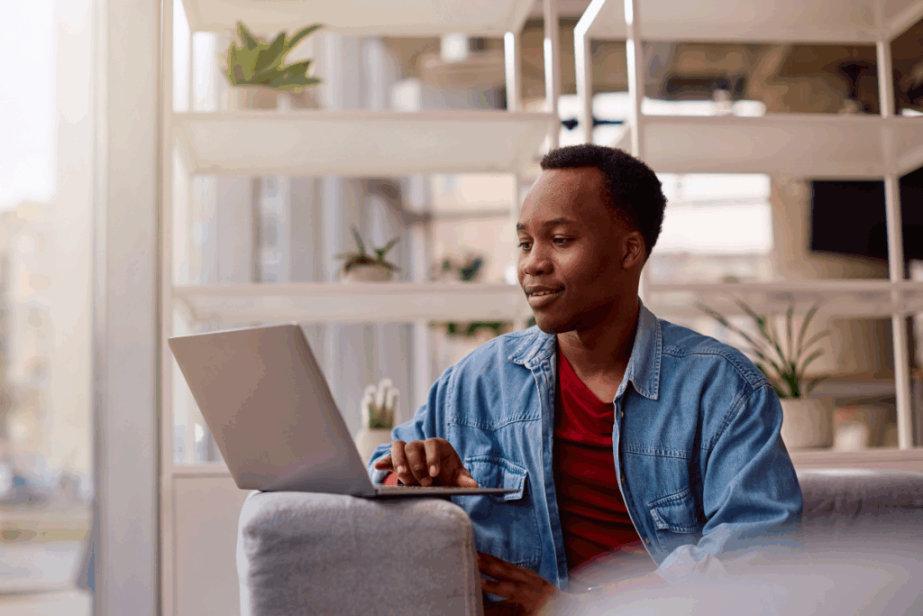 Um homem jovem negro está sentado em um ambiente interno iluminado, digitando em um notebook apoiado à sua frente