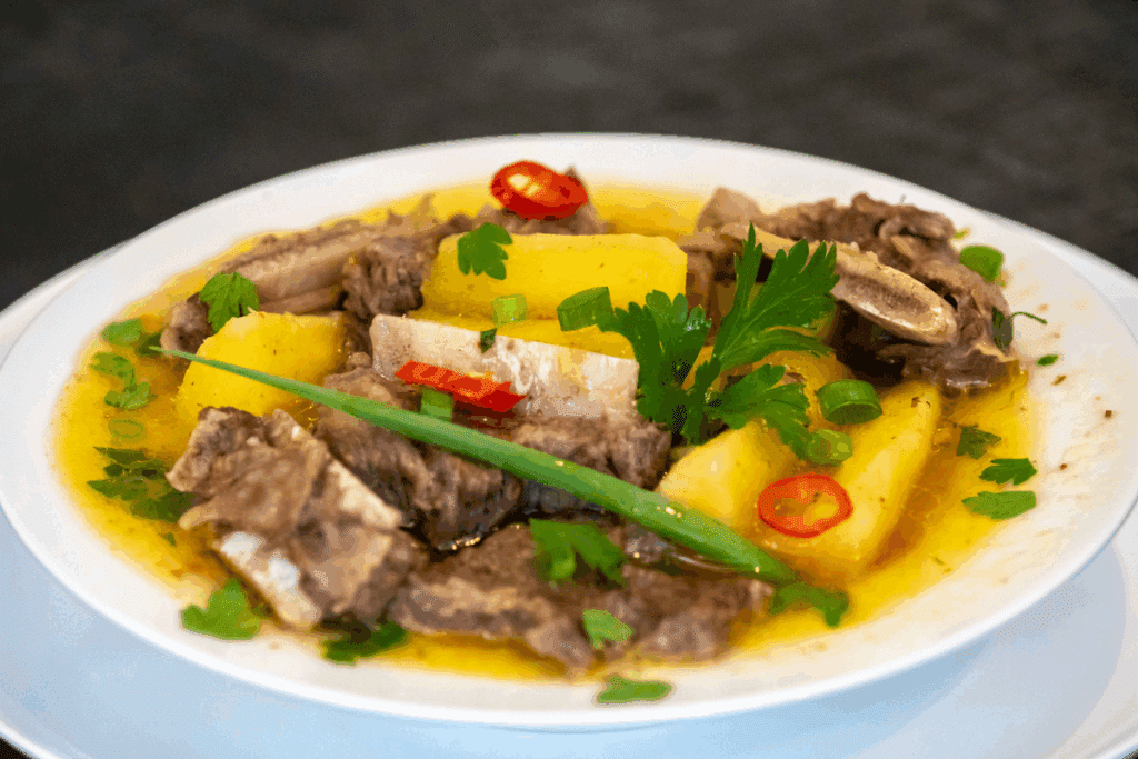 Close-up de uma porção de vaca atolada, com costela bovina, mandioca amarela e caldo encorpado, decorada com salsinha, rodelas de pimenta vermelha e um talo de cebolinha.